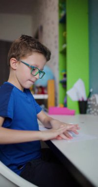 Gözlüklü genç bir çocuk beyaz bir masada oturuyor ve bir parça kağıt katlıyor. Gülümsüyor ve mavi bir tişört giyiyor..