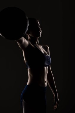 Silüete yakalanmış genç bir kadının dramatik bir yakın çekimi. Spor kıyafetleri içinde basketbol topunu zarif bir şekilde tutuyor..