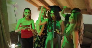 Heyecanlı bayan mezunlar mezuniyet günlerini bir mezuniyet partisinde kutluyor. Mezun, mezuniyet şapkası ve diplomasını göstererek arkadaşlarıyla selfie çektirmek için poz veriyor..
