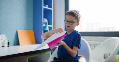 Gözlüklü genç bir çocuk odasında masasında oturmuş, pembe beyaz bir kağıt uçakla oynuyor..