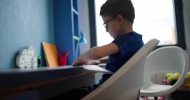 Gözlüklü genç bir çocuk masasında oturur ve dikkatle bir parça kağıt katlar. Odaklanmış ve faaliyetleriyle meşgul..