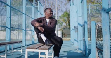 Mutlu bir adam padel kortunda bankta oturur, ayakkabılarını bağlar ve güneş gözlüğü takar, güneşli günün tadını çıkarır..