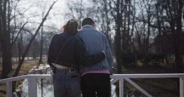 Genç bir çift parktaki beyaz bir köprüde duruyor. Huzurlu manzaranın tadını çıkarıyorlar..