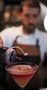 Bir barmen kalabalık bir barda renkli bir kokteyl hazırlar. Karışımın detaylarına dikkat ederken malzemeleri ustalıkla bir bardağa dolduruyor. Atmosfer canlı ve davetkar..
