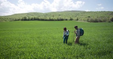 Güneşli bir günde, bulutlu, yeşil bir tarlada yürüyüş yapan çift. Sırt çantaları ve yürüyüş direkleri taşıyorlar..