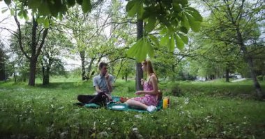 Ağaçlar ve yeşilliklerle çevrili parkta romantik bir pikniğin tadını çıkaran bir çift. Yiyecekleri, içecekleri ve sohbeti paylaşıyorlar. Barışçıl ve mutlu bir ortam yaratıyorlar..