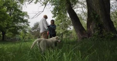 Couple enjoys a leisurely walk in the park with their dog, savoring the tranquility of nature, while holding a bowl and a red umbrella.
