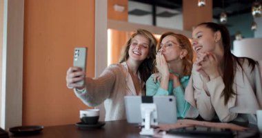 Üç iş kadını modern bir kafede birlikte vakit geçiriyorlar. Gülümsüyor ve selfie çekiyorlar, şık bir ortamda kahvelerini yudumlarken yoldaşlıklarını sergiliyorlar..