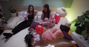 Bir grup kız, bir pijama partisi sırasında rahat bir oturma odasında balonlarla oynayıp birlikte eğlenirken içkilerini yudumluyorlar. Kahkaha ve arkadaşlık boşluğu doldurur..