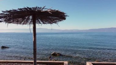 Güneşli bir günde geleneksel plaj şemsiyeleriyle Ohri Gölü manzarası. Sakin su ve uzak dağlar huzurlu bir atmosfer yaratır..