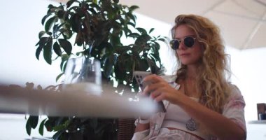 Güneşli bir öğleden sonra şık bir kadın bir kafede oturur. O, güneş gözlüğü takıyor, telefonuna odaklanmış, bir cam yanında oturuyor, yeşillik ile çevrili..