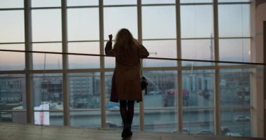 Kahverengi paltolu bir kadın modern bir binada büyük bir pencerenin önünde durmuş, şehri seyrediyor. Arkasını döndü ve telefonuyla konuşurken uzaklaştı..