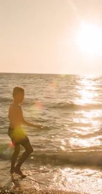 A young boys silhouette is captured as he playfully throws rocks into the calm ocean waves at sunset, with golden sunlight reflecting beautifully on the water during a peaceful summer evening on the