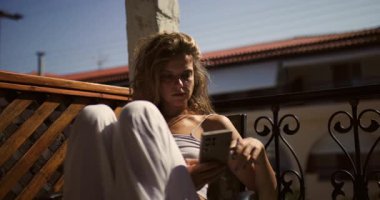 Bir kadın balkonunda huzurlu bir sabah geçirir. Telefonunda okurken güneş ışıl ışıl parlıyor. Güne rahat bir şekilde başlıyor..