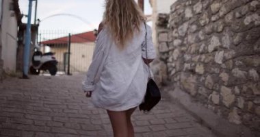 A person strolls peacefully down a narrow street lined with charming stone walls. The bright sun adds warmth to the lively atmosphere of the town.
