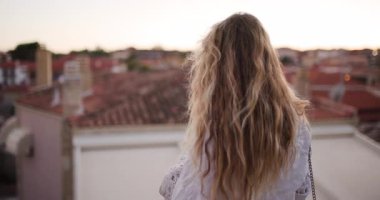 Çatıdan bakıldığında, uzun saçlı bir insan güneş batarken şehrin çatılarına bakıyor ve sıcak renklerle gökyüzünü aydınlatıyor. Bu huzurlu an, çevrenin güzelliğini yakalar..