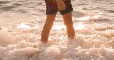 Golden light illuminates a persons bare legs as gentle ocean waves wash over their feet on a sandy beach during a beautiful sunset or sunrise.