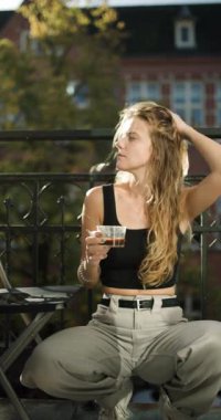 Bir kadın balkonda oturuyor, kahvesini yudumluyor ve öğleden sonra güneşinin tadını çıkarıyor. Rahatlamak ve çevresini takdir etmek için bir dakikasını ayırıyor. Huzurlu bir atmosfer yaratıyor..