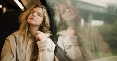 Dalgalı saçlı genç bir kadın, düşünceli bir şekilde tren penceresinden dışarı bakıyor. Kırsal kesim yanından geçiyor ve yansıttığı gibi sakin bir zemin oluşturuyor. İfadesi derin düşüncelere daldığını gösteriyor..