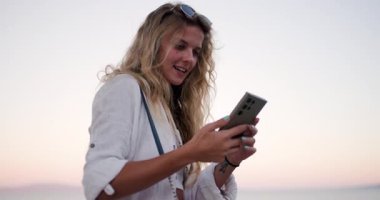 Uzun saçlı biri kumsalın yanında duruyor, güzel bir deniz manzarası büyülüyor. Gün batımında akıllı telefonlarını kullanarak gülümseyerek sakin atmosferi kucaklıyorlar..