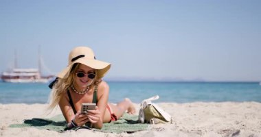 Güzel bir plaj gününde, bir kadın telefonuyla selfie çekerken gülümser. Şık bir şapka ve güneş gözlüğü takarak kumda dinlenirken güneş ışığı parlıyor..