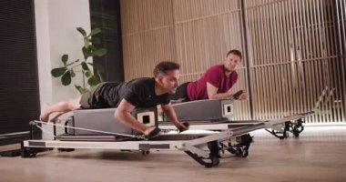 İki adam şık bir fitness stüdyosunda reformdan yararlanan odaklanmış bir egzersiz yapıyorlar. Atmosfer enerjiktir. Güç ve esnekliği hedef alan egzersizler sergilerler..