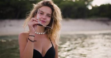 Genç bir kadın kıyıya yakın duruyor, kameraya gülümsüyor. Deniz kabuğundan bir kolye takıyor ve dalgalı saçları var. Sahilde sıcak, güneşli bir günün özünü yakalıyor..