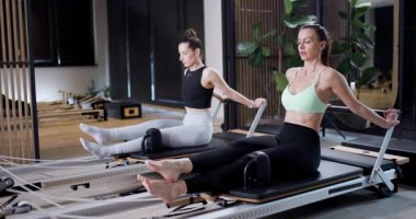İki kadın parlak bir stüdyoda yenilikçi makinelerle pilates egzersizi yapıyorlar. Atmosfer sakin, yeşil bitkiler ve ahşap dekorlar var. Hareketlerine ve şekillerine odaklanırlar..