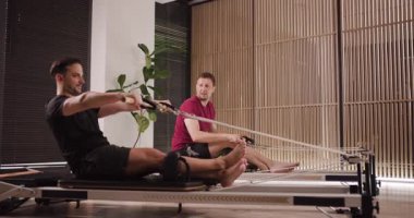İki adam bir hoş geldin fitness stüdyosunda pilates reformatörlüğü çalışmasına katılıyor. Güç ve duruşa odaklanırlar. Egzersiz için enerji verici bir atmosfer yaratırlar..