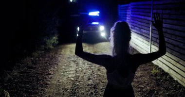 Bir kız elleri havada, ağaçlarla çevrili karanlık bir yolda teslim olmuş vaziyette dikiliyor. Yanıp sönen ışıkları olan bir polis aracı yaklaşıyor ve geceleri yoğun bir atmosfer yaratıyor..