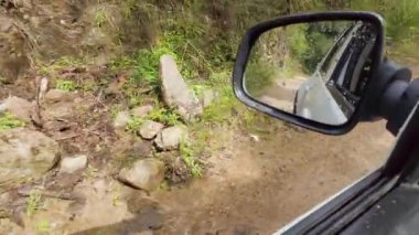 Bir araba, açık mavi gökyüzünün altında canlı bitkiler ve ağaçlarla kaplı dolambaçlı toprak bir yolda ilerliyor. Sürücü bölgeyi keşfederken doğanın sakinleştirici atmosferinin tadını çıkarıyor. Su sıçraması.