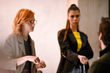 İki kadın, modern bir ofiste fikir ve içgörü paylaşımı yaparak bir tartışmaya girişirler. Bir kadın gözlük ve spor ceket giyerken, diğeri şık bir kıyafet giyiyor..