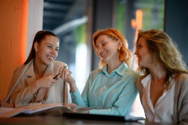 Üç gülümseyen kadın modern ofis ortamında bir masanın etrafında oturuyor, muhtemelen iş tartışıyorlar ve öğleden sonra bir toplantı sırasında neşeli bir anı paylaşıyorlar..