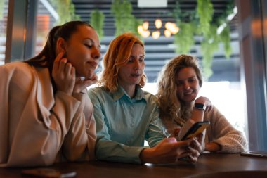 Üç iş kadını aynı fikirleri paylaşıyor ve şık bir kafede birbirlerine eşlik etmekten keyif alıyorlar, canlı bir atmosferde projeler üzerinde çalışırken telefonlarına odaklanıyorlar..