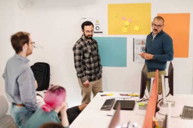 Bir grup iş adamı, çağdaş bir ofiste beyin fırtınası yapıyor. Fikirlerini paylaşıyorlar ve bilgisayarlar etrafında işbirliği yapıyorlar, duvarda renkli notlar sergileniyor..