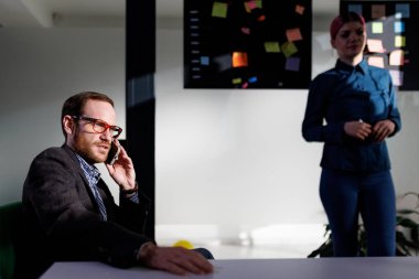 Bir işadamı bir telefon görüşmesi masasında otururken, diğeri yakınlarda duruyor, hem üretkenlik ve işbirliği odaklı, parlak, organize bir ofiste..