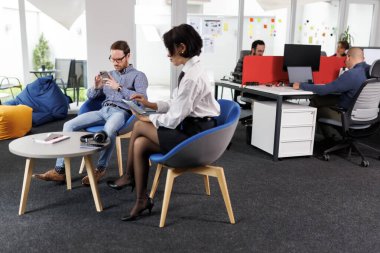 Modern ofis ortamında iki iş adamı tartışıyorlar. Bir kişi bir cihaz üzerinde kaydırırken, diğeri bir tablet kullanıyor. Ekip üyeleri yakınlardaki masalarda çalışıyor..