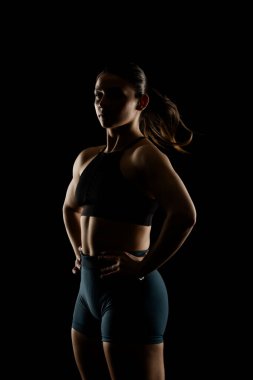 Atletik giyinen güçlü ve kendine güvenen bir kadın karanlık bir arka plana karşı poz verir. Zindelik, güç ve güçlendirme kavramları için mükemmel..