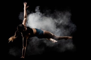 Dinamik yoga pozu veren bayan sporcu dumanlı bir bulutun ortasında, simsiyah bir arkaplan üzerine kurulmuş, somut güç, denge ve konsantrasyon..