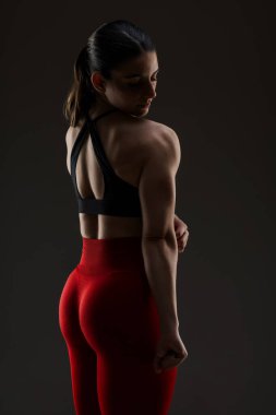 Kırmızı taytlı ve siyah spor sutyenli formda bir kadın sırt ve omuz kaslarını gösteriyor. Güçlü duruşu, zindeliğini ve gücünü vurguluyor..