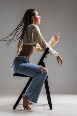 Bir kadın zarif bir şekilde sandalyeye oturur, örgülü saçları sırtına doğru dökülür. Modaya uygun kesilmiş bir kazak ve yırtık kot pantolon giyerken kendine güveni tam..
