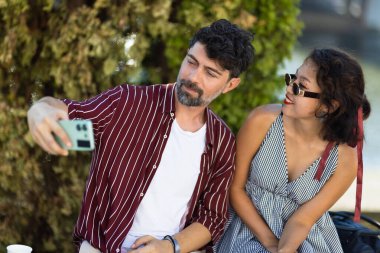 Güneşli bir günde dışarıda selfie çeken genç bir çift. Mutlulukları ve günlük hayatları, rahatlatıcı bir yaz atmosferinde dostluk ve rahatlığı yansıtıyor..