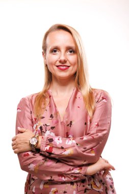 İş kadını, çiçekli bir bluz giyerek kollarını kavuşturarak güvenle ayakta duruyor. Dostane bir gülümseme sergiliyor. Parlak bir stüdyo ortamında profesyonelliği somutlaştırıyor..