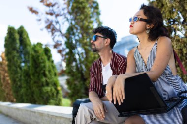 Güneş gözlüklü iki şık şahıs parkta bir bankta oturuyor, dizüstü bilgisayarla uğraşıyorlar, kentsel yaşam tarzını ve doğayı kucaklıyorlar..