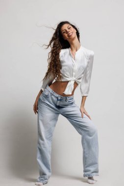 Beyaz bir bluz ve kot pantolon giymiş, dinamik bir poz veren, kendine güveni olan gülümseyen bir kadın.