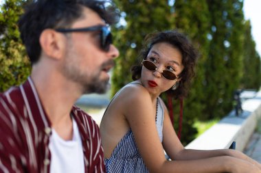 Güneş gözlüklü ve kırmızı rujlu bir kadın, güneşli bir günde ön planda bulanık bir adamla otururken şaşkınlıkla bakıyor..