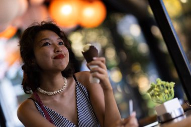 Genç bir kadın, sıcacık bir kafede dondurma külahının tadını çıkarıyor. Sıcak, ortam aydınlatmasıyla dolu olan çevreye hayranlıkla gülümsüyor..