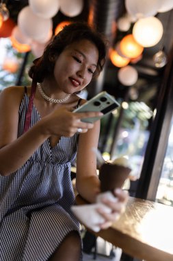 Genç bir kadın, şık bir kafede, sıcacık, sıcacık bir aydınlatmayla çevrili akıllı bir telefonla dondurmasının tadını çıkarıyor..