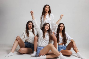 Dost canlısı ve neşeli bir atmosferde birlikte poz veren dört genç kadın, minimalist beyaz bir stüdyo ortamında aralarındaki bağı ve pozitif enerjiyi gösteriyorlar..