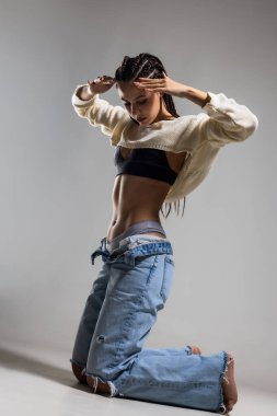 Örgülü güzel bir kadın sıradan bir ortamda diz çöker, örülmüş bir bluz ve sıkıntılı bir kot pantolon giyer. Kendine güvenen bir duruş sergiliyor, moda anlayışını ve zarafetini vurguluyor..